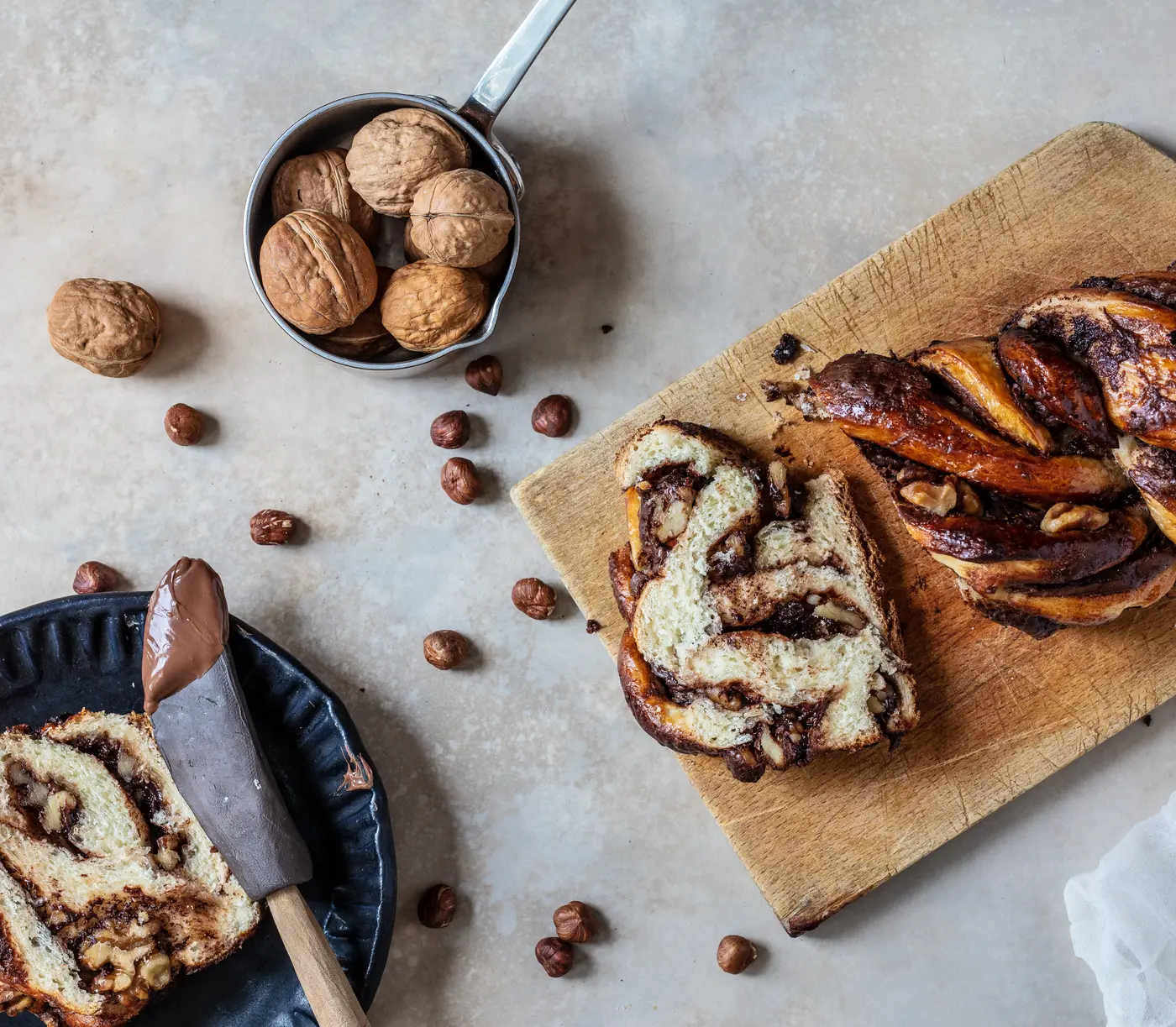 Chocolate babka loaf on a wooden board with two cut slices showing the chocolate swirl, surrounded by walnuts and hazelnuts.