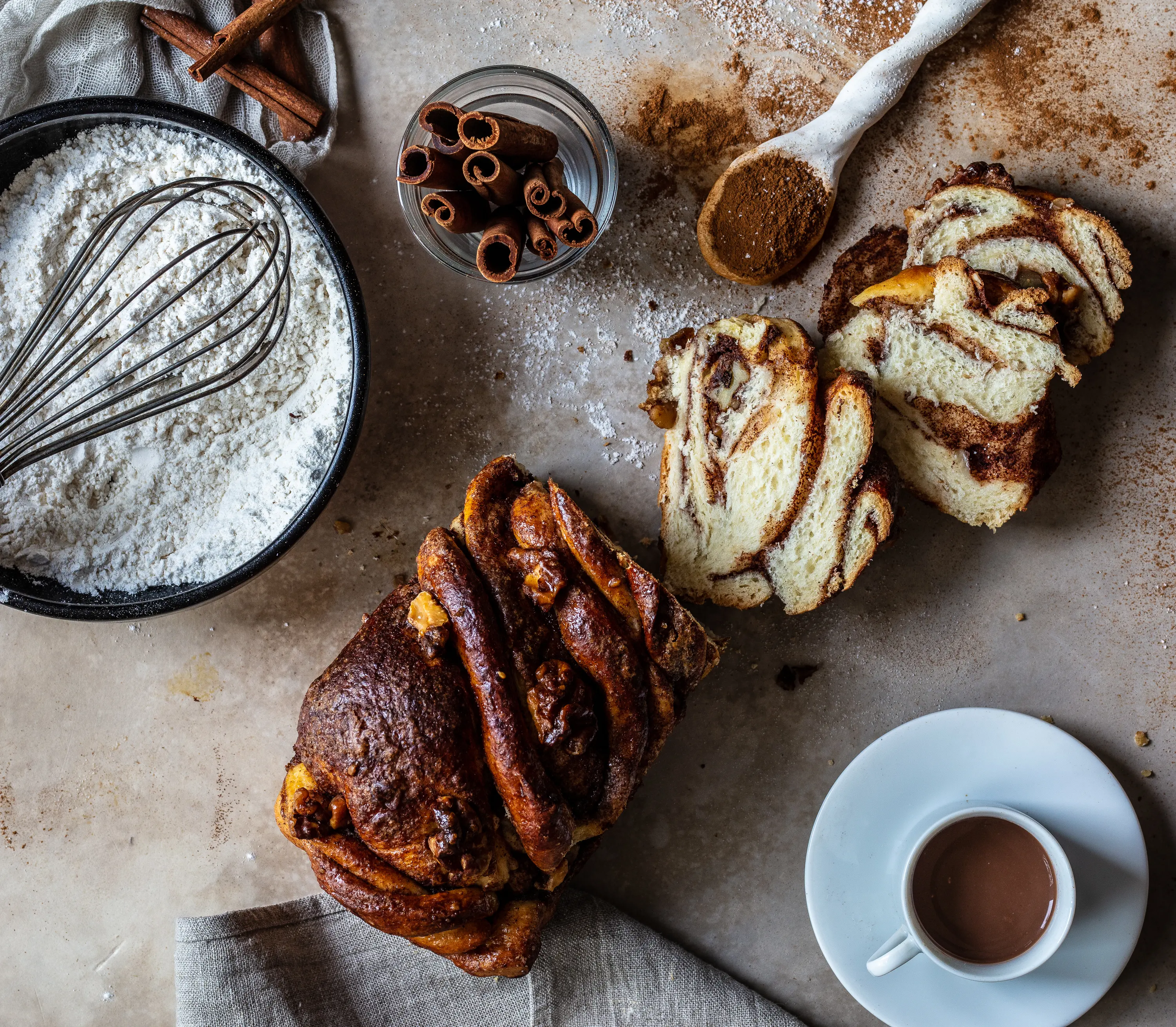 Cinnamon babka, whole loaf and cut slices, with flour, cinnamon sticks and a cup of coffee.