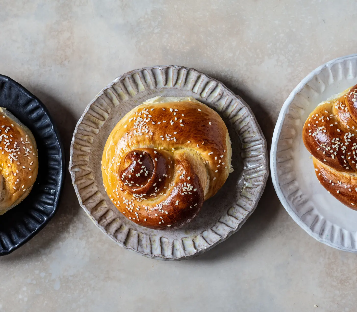 Three sesame challah knots on ceramic plates in black, grey and white.