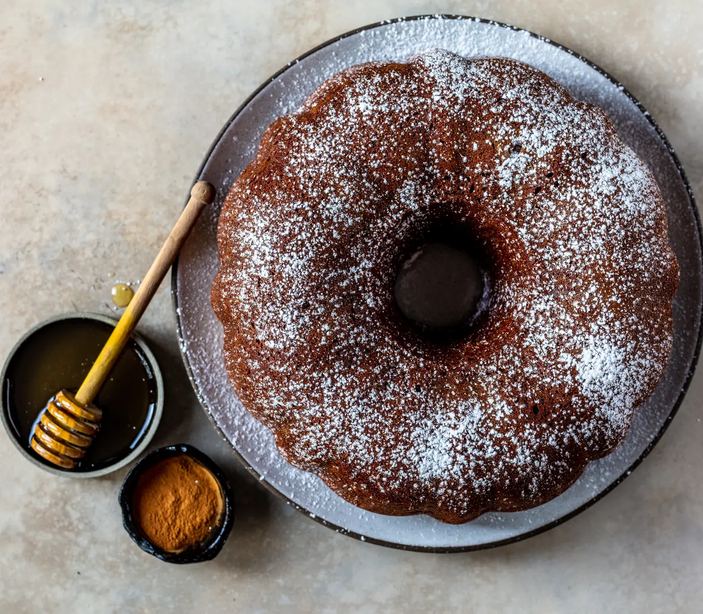 A ring-shaped honey cake dusted with powdered sugar on a ceramic plate, with a dish of honey and a honey dipper.